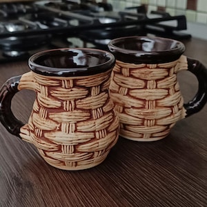 May include: Two brown ceramic mugs with a woven pattern. The mugs have brown handles and are sitting on a wooden surface.