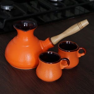 May include: An orange ceramic coffee pot with a wooden handle and two matching small coffee cups. The interior of the pot and cups are dark brown. The set is on a dark brown surface.