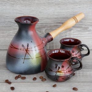 May include: A ceramic coffee pot with a long wooden handle and two matching mugs, all featuring a gradient finish of red, black, and yellow with hand-painted dragonfly motifs. Several roasted coffee beans are scattered around the base of the set.