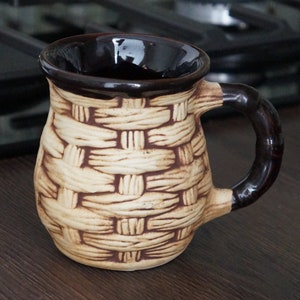 May include: A brown ceramic mug with a woven pattern and a brown handle. The mug is sitting on a dark brown surface.