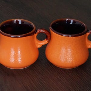 May include: Two orange ceramic mugs with brown rims and handles. The interior of each mug is dark brown. The mugs have a rounded shape and sit on a dark wooden surface. The mugs are side by side.
