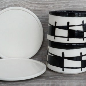 May include: Two stacked white ceramic mugs with black geometric designs and matching white saucers. The mugs have black handles and a black rim. The saucers are round and have a slightly raised edge. The background is a light wood grain.