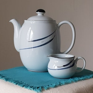 Può includere: Un set da tè composto da teiera e lattiera bianca con un design astratto blu e nero. La teiera ha un coperchio e un manico. La lattiera ha un manico. Il set è appoggiato su un sottopentola blu turchese.