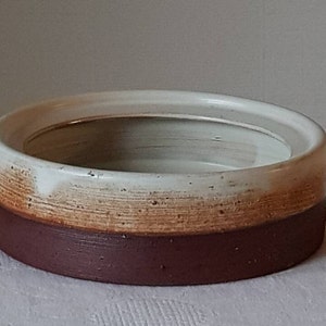 Op de afbeelding: Een ronde keramische kom met een witte glazuur en een bruine en oranje glazuurband. De kom heeft een licht afgeronde rand.