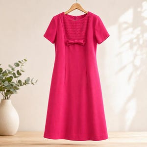 Può includere: Un abito fucsia brillante al ginocchio con maniche corte, scollo rotondo e un pannello del corpetto arricciato. Un piccolo fiocco decorativo è centrato appena sotto la sezione arricciata e la gonna cade in una linea dritta.