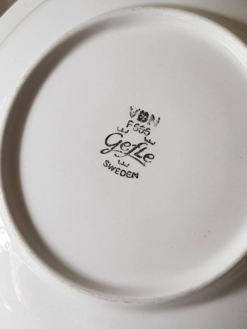 以下が含まれることがあります： 「VDN F555 Gefle SWEDEN」と書かれた黒いスタンプが付いた白いセラミックプレート。プレートにはわずかに盛り上がった縁と滑らかな表面があります。スタンプはプレートの中央にあります。