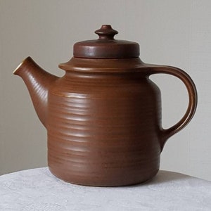 Könnte beinhalten: Eine braune Teekanne aus Keramik mit Deckel und Henkel. Die Teekanne hat eine strukturierte Oberfläche und eine leicht abgerundete Form.