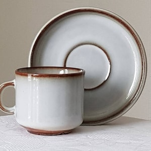 以下が含まれることがあります： 白のセラミックコーヒーカップとソーサーのセット。カップは丸みを帯びた形で、小さなハンドルが付いています。カップの縁と底、ソーサーの縁は温かみのある茶色です。セットは白い表面にあります。