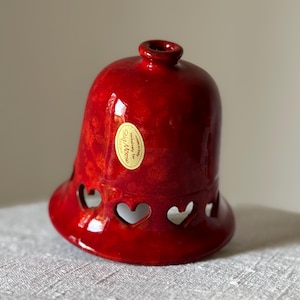 May include: A glossy red ceramic bell-shaped candle holder with heart-shaped cutouts. The bell has a flared base and a small opening at the top. A gold sticker is affixed to the side. The candle holder is on a neutral surface.