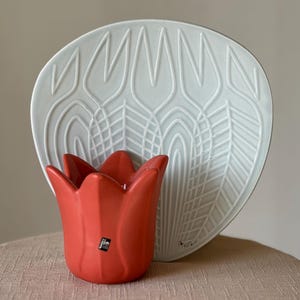 May include: A red tulip-shaped candle holder with a white decorative plate in the background. The plate has a floral design. The candle holder and plate are on a light brown surface.