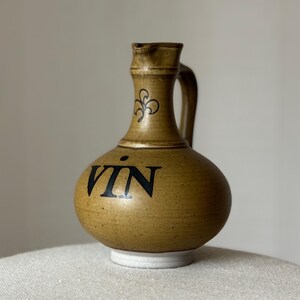 Op de afbeelding: Een bruine keramische wijnkan met een zwarte inscriptie die "VIN" zegt. De kan heeft een handvat en een decoratief ontwerp op de hals.