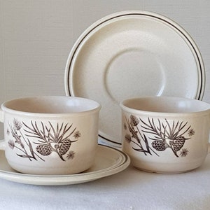 以下が含まれることがあります： 茶色の松ぼっくり模様の白いティーカップ2つと、それに合うソーサー。カップとソーサーはクリーム色の背景です