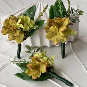 May include: Three buttonhole arrangements with yellow and green flowers, dark green stems and leaves. The flower petals have a hint of pink. The stems are wrapped in dark green ribbon. Set against a white textured background.