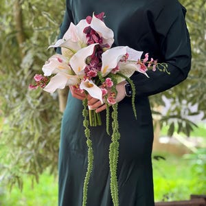 May include: A floral bouquet featuring white calla lilies, deep burgundy orchids, and long, trailing green amaranth stems. The arrangement is held together by a wrapped stem base, showcasing a contrast between the white petals and dark foliage.