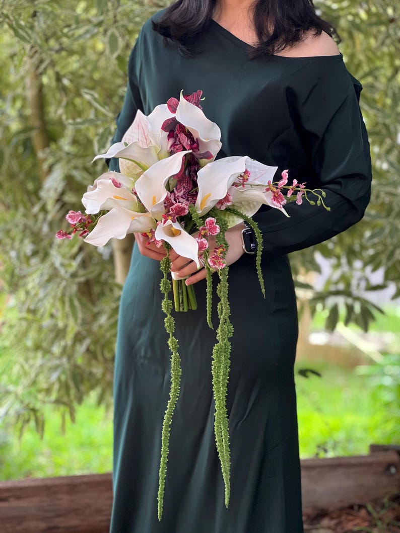 May include: A floral bouquet featuring white calla lilies, deep burgundy orchids, and long, trailing green amaranth stems. The arrangement is held together by a wrapped stem base, showcasing a contrast between the white petals and dark foliage.