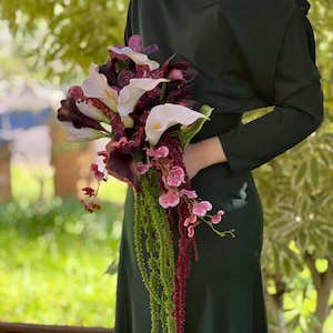 May include: A floral bouquet featuring white and dark purple calla lilies, deep burgundy orchids with speckled patterns, and long, trailing green and dark red amaranth stems. The arrangement is held against a solid dark green long-sleeve dress.