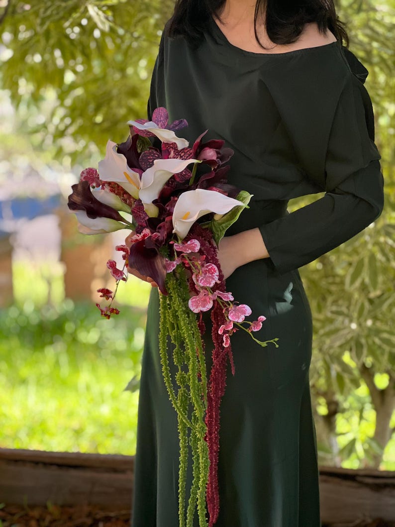 May include: A floral bouquet featuring white and dark purple calla lilies, deep burgundy orchids with speckled patterns, and long, trailing green and dark red amaranth stems. The arrangement is held against a solid dark green long-sleeve dress.