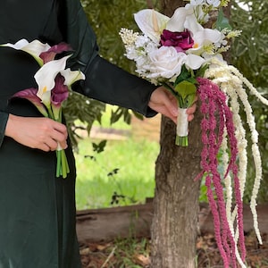 Pu&ograve; includere: Due mazzi di fiori. Un mazzo presenta calle bianche con centri viola scuro. L'altro mazzo include rose bianche, fiori bordeaux e amaranto bordeaux e bianco. I fiori sono tenuti da una persona che indossa un abito verde scuro.