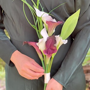 Pu&ograve; includere: Un bouquet di calle con fiori bianchi e bordeaux scuro, steli verdi e lunghi accenti verdi simili all'erba. I fiori sono legati insieme con un nastro bianco. Lo sfondo &egrave; un vestito verde scuro.