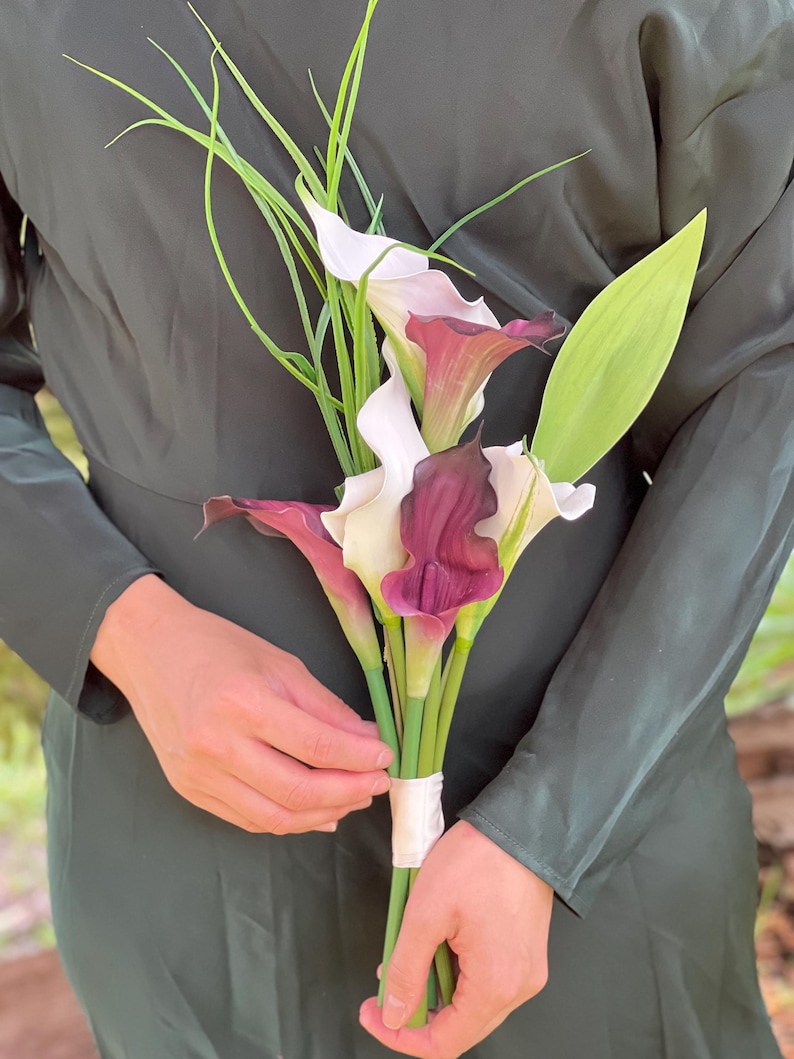 Pu&ograve; includere: Un bouquet di calle con fiori bianchi e bordeaux scuro, steli verdi e lunghi accenti verdi simili all'erba. I fiori sono legati insieme con un nastro bianco. Lo sfondo &egrave; un vestito verde scuro.
