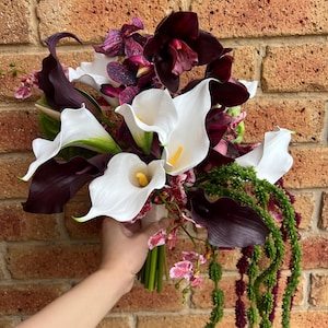 May include: A floral bouquet featuring white calla lilies with yellow spadices, deep purple calla lilies, dark maroon orchids with purple speckles, and long, trailing green amaranth strands. The flowers are gathered together in a bundle.