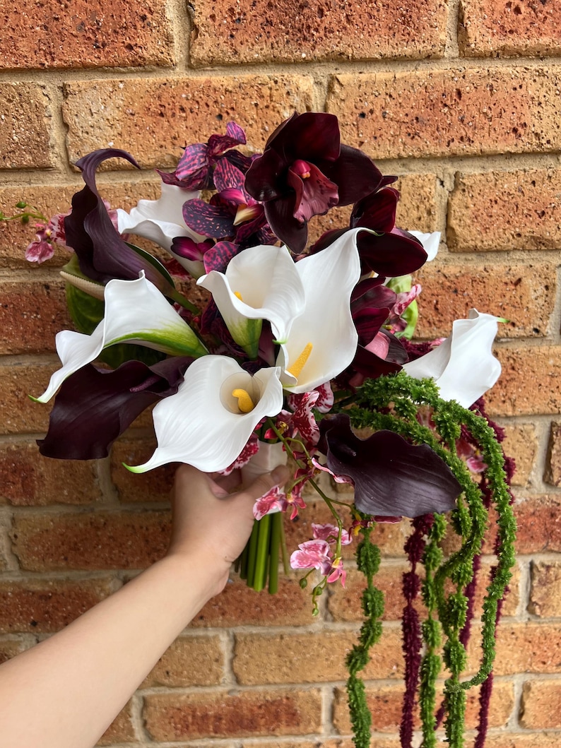 May include: A floral bouquet featuring white calla lilies with yellow spadices, deep purple calla lilies, dark maroon orchids with purple speckles, and long, trailing green amaranth strands. The flowers are gathered together in a bundle.