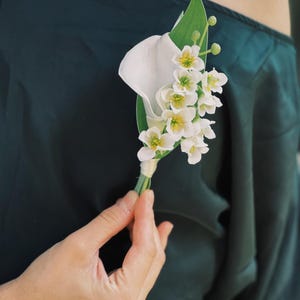 Pu&ograve; includere: Un fiore all'occhiello composto da una calla bianca e piccoli fiori bianchi con centri gialli, accentuati da foglie e steli verdi. La composizione floreale &egrave; tenuta contro un capo di abbigliamento verde scuro.