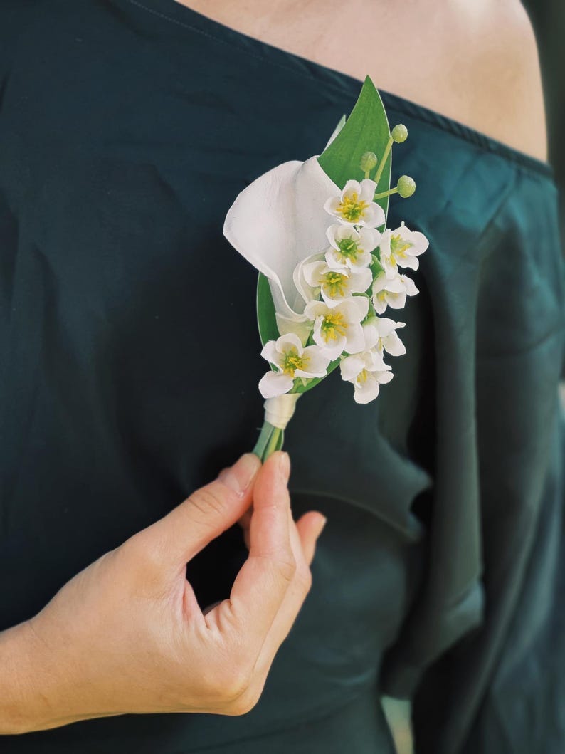 Pu&ograve; includere: Un fiore all'occhiello composto da una calla bianca e piccoli fiori bianchi con centri gialli, accentuati da foglie e steli verdi. La composizione floreale &egrave; tenuta contro un capo di abbigliamento verde scuro.