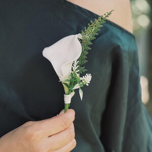 Pu&ograve; includere: Un'occhiello di calla bianca con fogliame verde e piccoli fiori bianchi, avvolto con un nastro bianco. L'occhiello &egrave; tenuto contro un tessuto verde scuro.