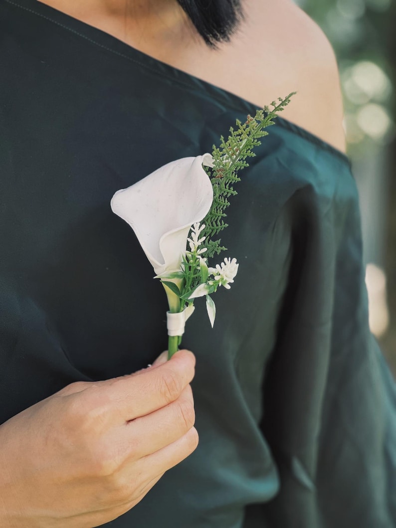 Pu&ograve; includere: Un'occhiello di calla bianca con fogliame verde e piccoli fiori bianchi, avvolto con un nastro bianco. L'occhiello &egrave; tenuto contro un tessuto verde scuro.