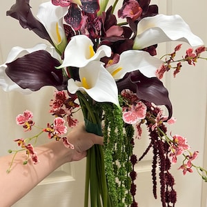 May include: A floral bouquet featuring white and dark purple calla lilies, deep burgundy orchids with speckled patterns, and smaller pink and white mottled orchids. The arrangement includes long green and dark red hanging amaranth stems.