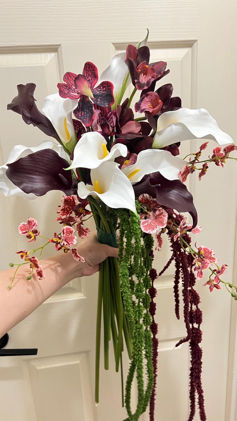 May include: A floral bouquet featuring white and dark purple calla lilies, deep burgundy orchids with speckled patterns, and smaller pink and white mottled orchids. The arrangement includes long green and dark red hanging amaranth stems.