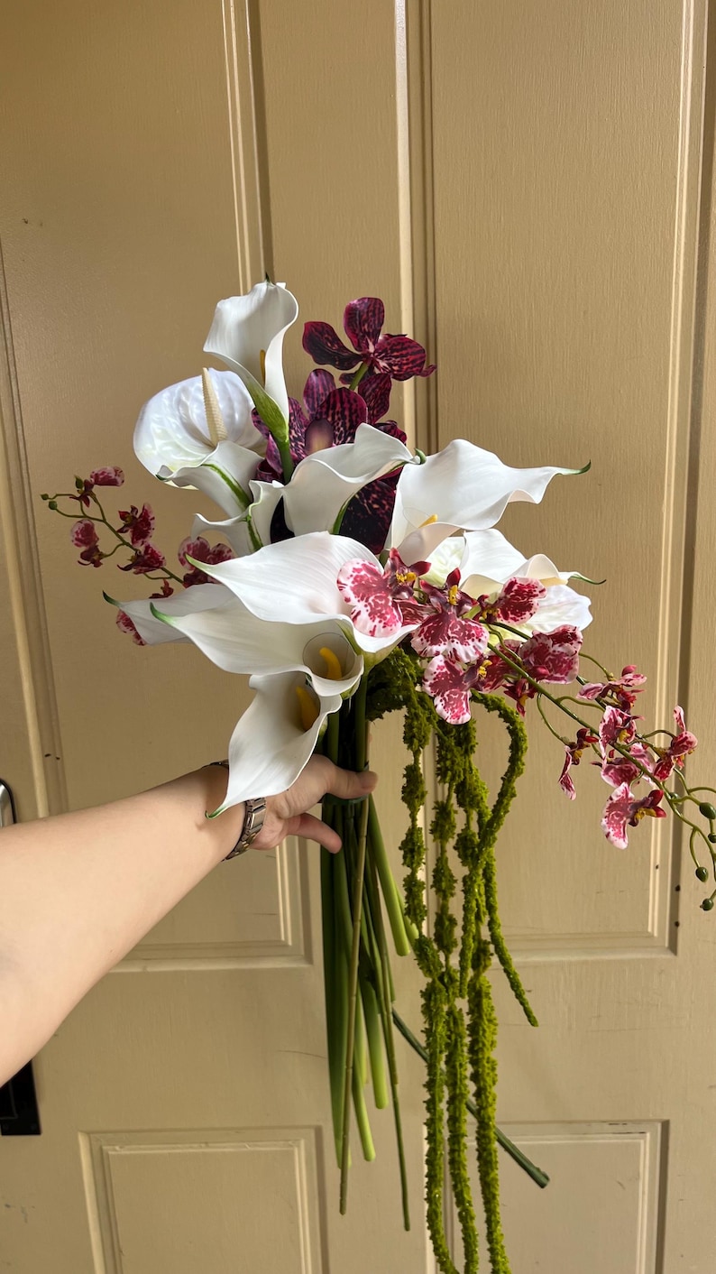 May include: A floral bouquet featuring white calla lilies, dark purple and white speckled orchids, and long, trailing green amaranthus stems. The arrangement is gathered into a bundle with visible green stalks extending downward from the flower heads.