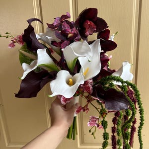 May include: A floral bouquet featuring white calla lilies with yellow spadices, deep burgundy calla lilies, dark purple cymbidium orchids, and speckled purple and white orchids, accented with long, trailing green and dark red amaranth stems.