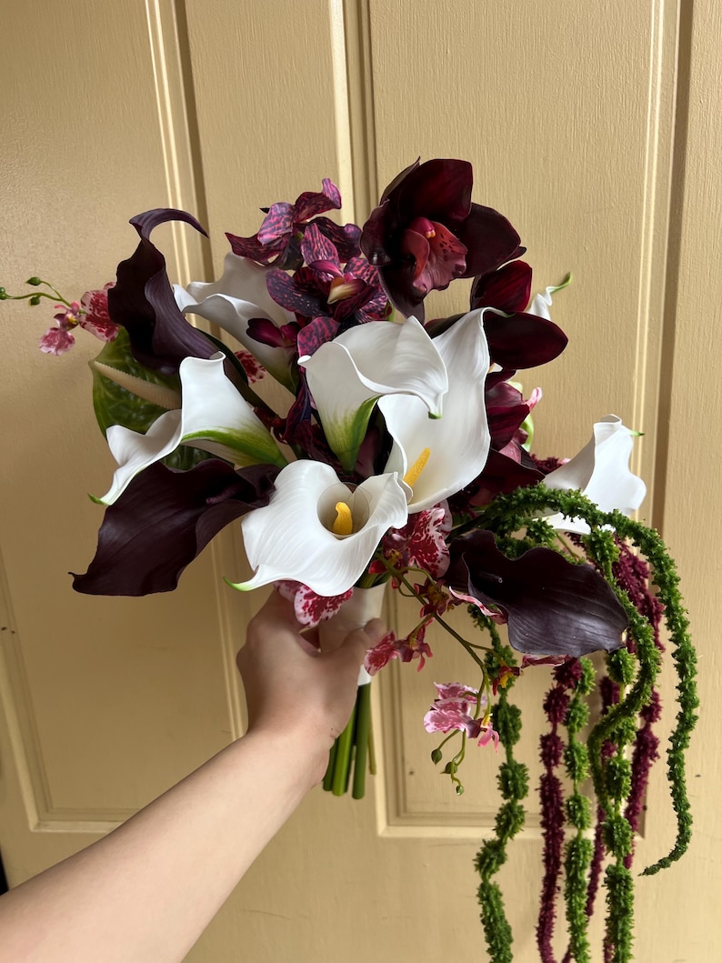 May include: A floral bouquet featuring white calla lilies with yellow spadices, deep burgundy calla lilies, dark purple cymbidium orchids, and speckled purple and white orchids, accented with long, trailing green and dark red amaranth stems.