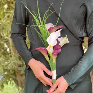 Pu&ograve; includere: Un bouquet di calle con petali bianchi e viola scuro, accentuato da un fogliame verde simile all'erba. I fiori sono tenuti insieme da un nastro color crema. Lo sfondo &egrave; un vestito verde scuro.