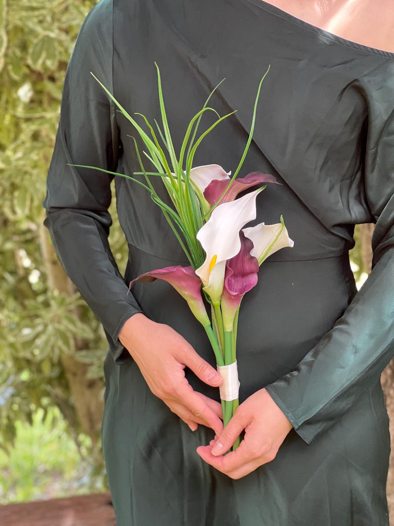Pu&ograve; includere: Un bouquet di calle con petali bianchi e viola scuro, accentuato da un fogliame verde simile all'erba. I fiori sono tenuti insieme da un nastro color crema. Lo sfondo &egrave; un vestito verde scuro.