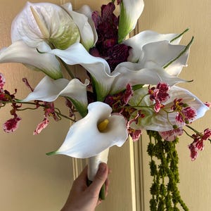 May include: A floral bouquet featuring large white calla lilies, a prominent white anthurium with a pale spadix, and deep purple speckled orchids. The arrangement includes cascading green amaranthus and is bound at the base with white ribbon.