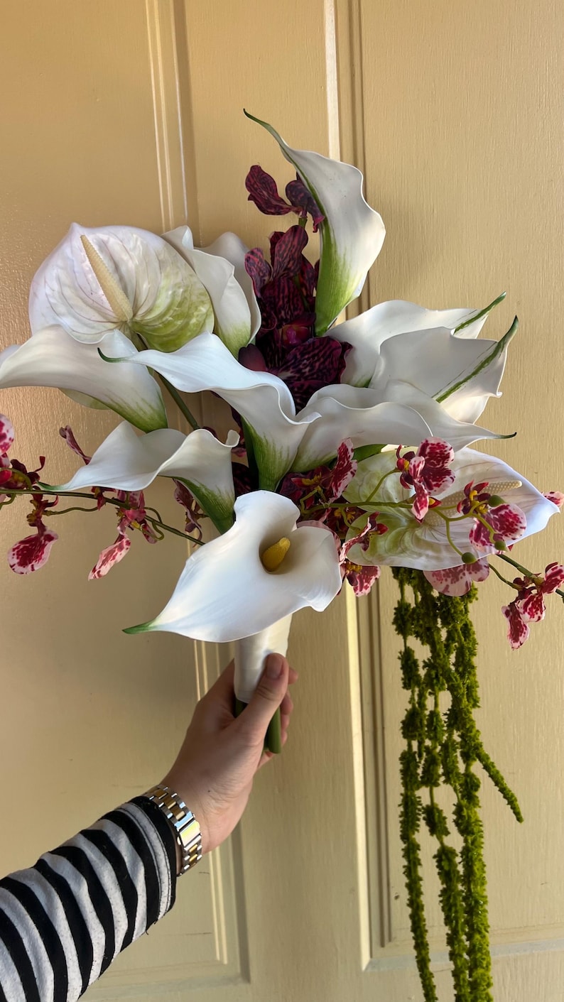 May include: A floral bouquet featuring large white calla lilies, a prominent white anthurium with a pale spadix, and deep purple speckled orchids. The arrangement includes cascading green amaranthus and is bound at the base with white ribbon.
