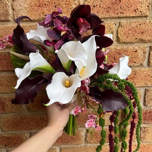 May include: A floral bouquet featuring white calla lilies with yellow centers, deep burgundy orchids, and dark purple calla lilies. The arrangement includes long, trailing green amaranth stems and small pink and purple speckled orchid blossoms.