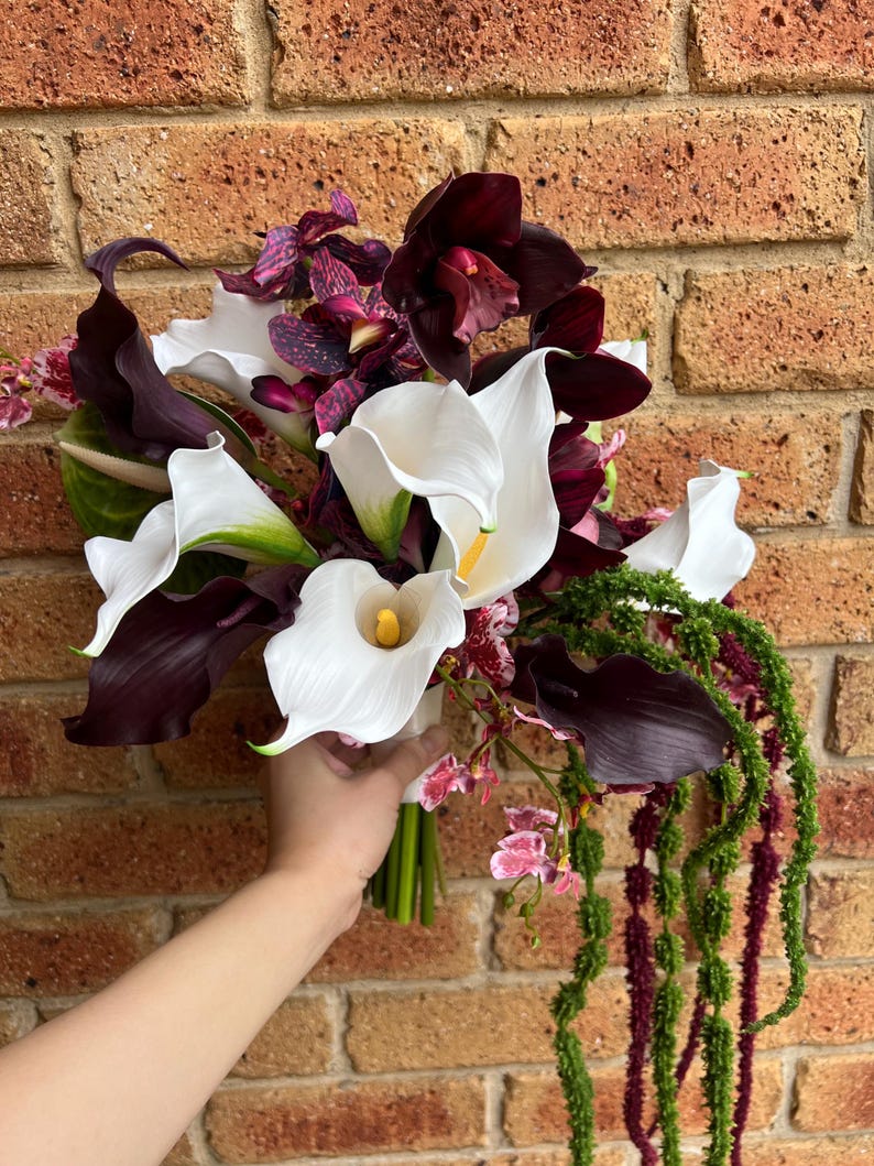 May include: A floral bouquet featuring white calla lilies with yellow centers, deep burgundy orchids, and dark purple calla lilies. The arrangement includes long, trailing green amaranth stems and small pink and purple speckled orchid blossoms.