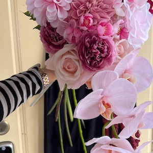 May include: A vibrant bouquet of pink and purple flowers, including peonies, roses, and orchids. The arrangement features various shades of pink and purple, with green stems and a dark red accent.