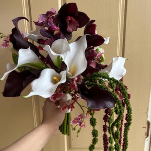 May include: A floral bouquet featuring white calla lilies with yellow centers, deep burgundy calla lilies, dark purple orchids with speckled patterns, and long, trailing green and maroon amaranth stems tied together at the base with a white ribbon.