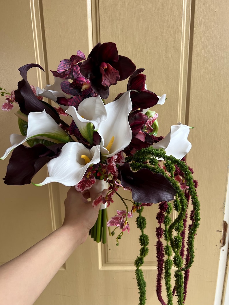 May include: A floral bouquet featuring white calla lilies with yellow centers, deep burgundy calla lilies, dark purple orchids with speckled patterns, and long, trailing green and maroon amaranth stems tied together at the base with a white ribbon.