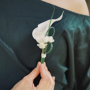 Pu&ograve; includere: Un'asola di fiori di calla bianchi e piccoli fiori bianchi con accenti verdi. La composizione floreale &egrave; tenuta da una persona che indossa un abito verde scuro. L'asola &egrave; avvolta con un nastro bianco.