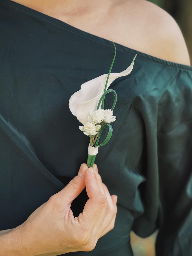 Pu&ograve; includere: Un'asola di fiori di calla bianchi e piccoli fiori bianchi con accenti verdi. La composizione floreale &egrave; tenuta da una persona che indossa un abito verde scuro. L'asola &egrave; avvolta con un nastro bianco.