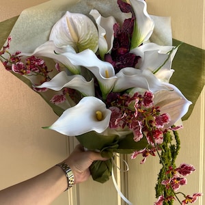 May include: A floral bouquet featuring white calla lilies with yellow spadices, white anthuriums, and dark red and white speckled orchids. The arrangement is wrapped in translucent paper and green foliage, with a long, trailing green stem.