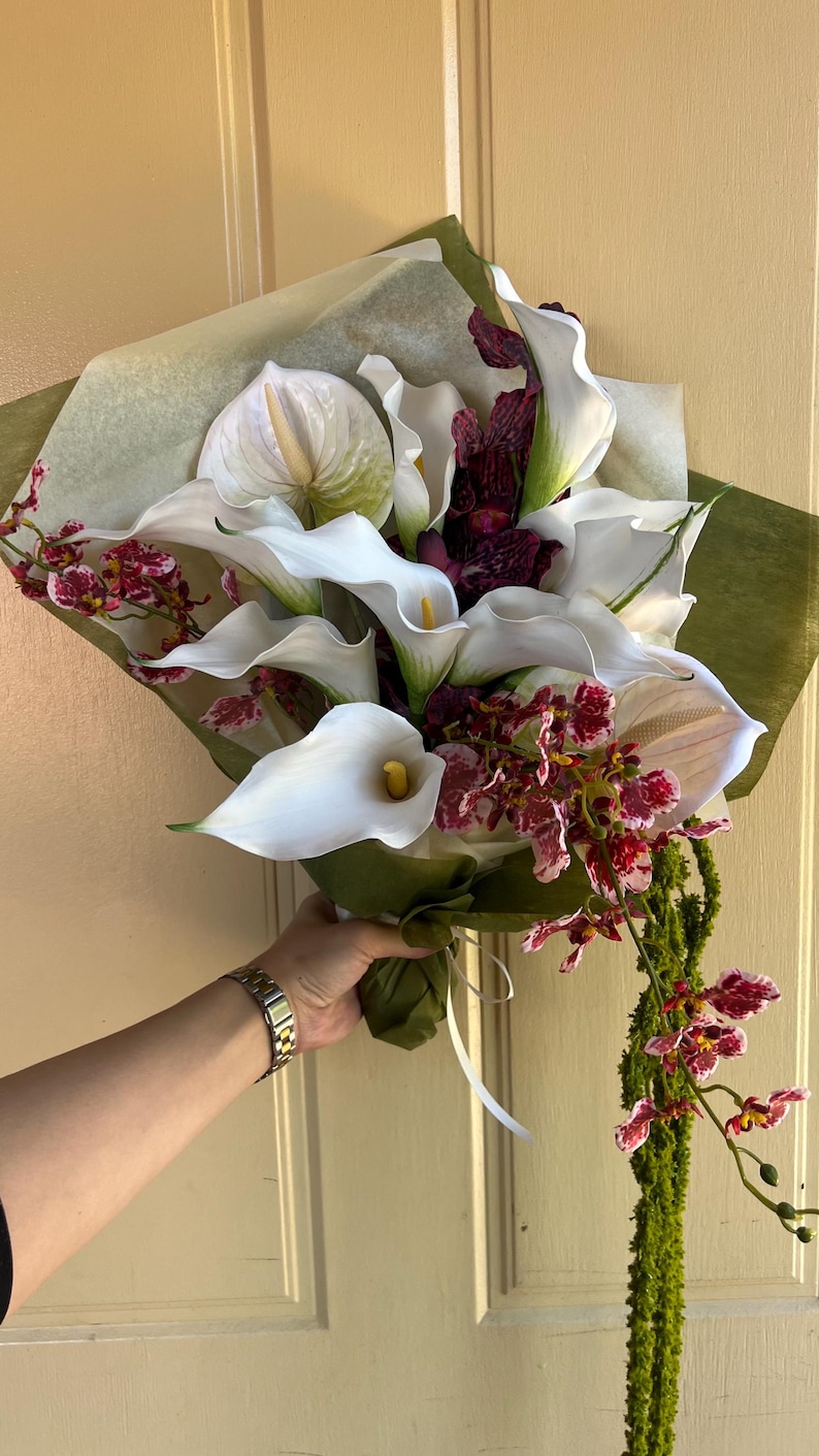 May include: A floral bouquet featuring white calla lilies with yellow spadices, white anthuriums, and dark red and white speckled orchids. The arrangement is wrapped in translucent paper and green foliage, with a long, trailing green stem.