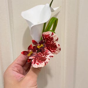 May include: A floral boutonniere featuring a single white calla lily paired with two smaller, mottled red and white orchid blossoms. The arrangement includes a slender green leaf accent, creating a compact and structured botanical display.