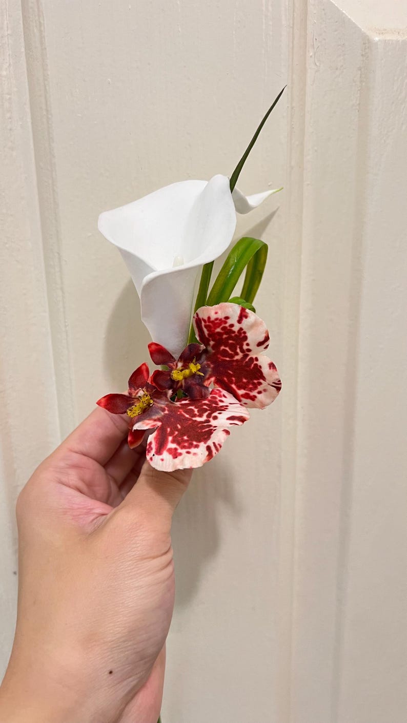 May include: A floral boutonniere featuring a single white calla lily paired with two smaller, mottled red and white orchid blossoms. The arrangement includes a slender green leaf accent, creating a compact and structured botanical display.